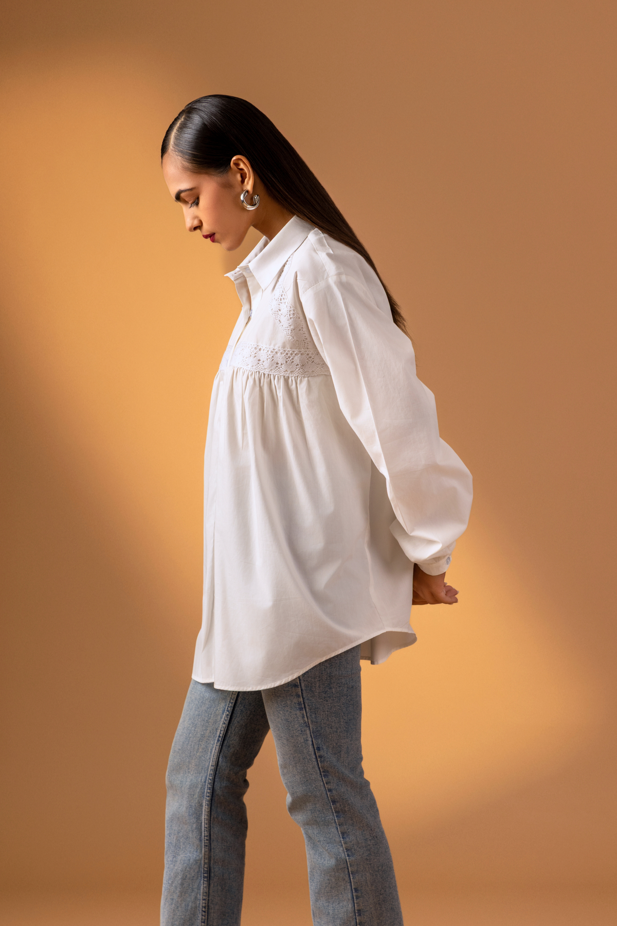 Side view of a model wearing a white button-down shirt with lace detailing, paired with blue jeans, set against a warm beige backdrop.
