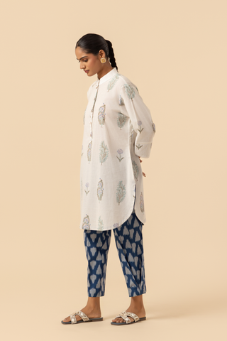 Side view of a woman stands wearing a light-colored, floral-printed kurta paired with blue patterned pants and sandals, against a beige background.