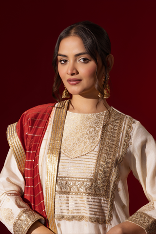 Closeup view of a woman in a white traditional outfit with gold embellishments and a red dupatta, posed against a rich red background.