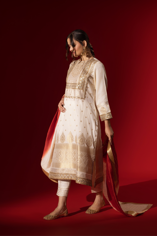 Side view of a woman in a white traditional outfit with gold embellishments and a red dupatta, posed against a rich red background.