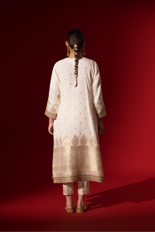 Back view of a woman in a white traditional outfit with gold embellishments and a red dupatta, posed against a rich red background.