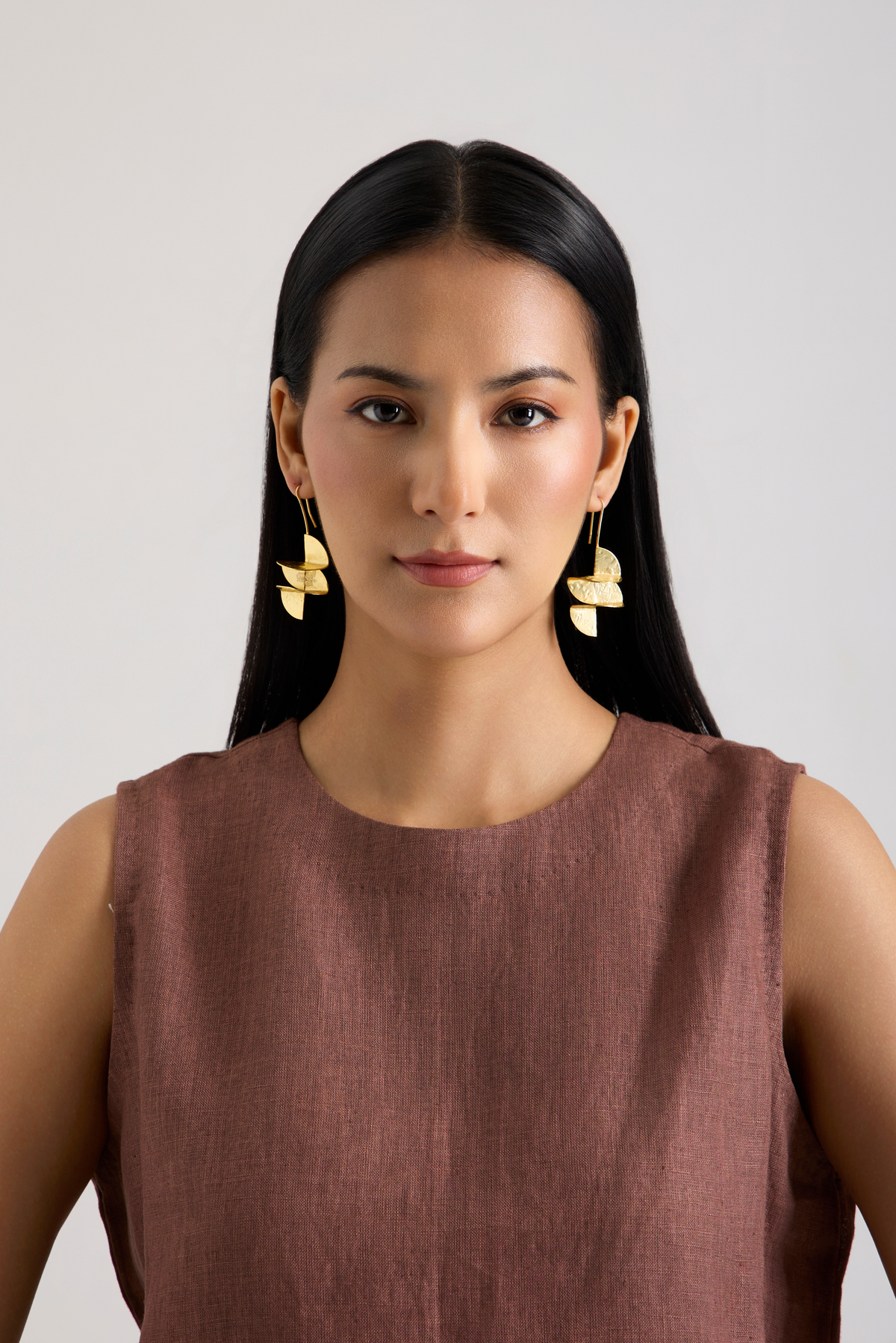 A woman with long black hair wearing gold leaf-shaped dangle earrings and a sleeveless brown top, photographed against a plain light gray background.