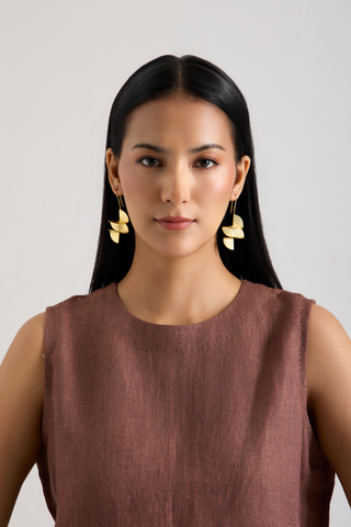 A woman with long black hair wearing gold leaf-shaped dangle earrings and a sleeveless brown top, photographed against a plain light gray background.