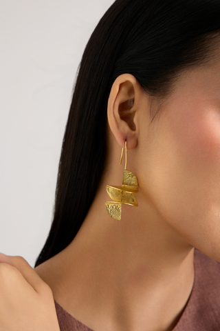 A close-up of a model's ear wearing a textured gold dangle earring with layered organic shapes, set against a neutral light gray background.