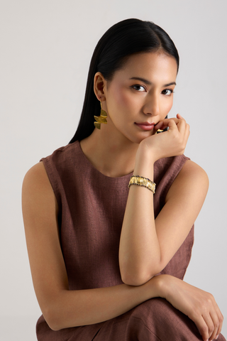 A woman with long black hair posing in a sleeveless brown dress, wearing a gold link bracelet and matching gold earrings against a light gray background.