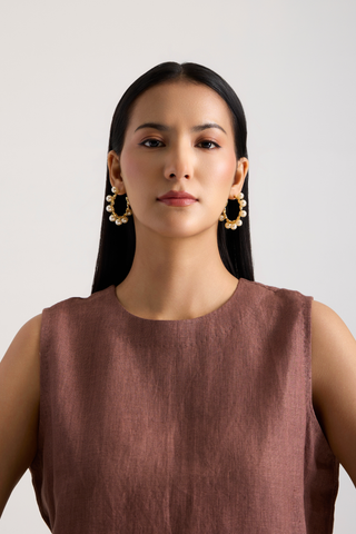 A woman with long black hair is shown from the chest up against a light gray studio background, wearing large gold-toned hoop earrings with clear stones and a sleeveless mauve-brown top.