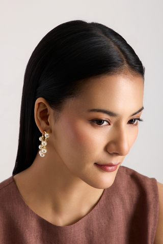 A close-up portrait of a woman with dark hair wearing a gold and pearl drop earring, set against a plain light gray background.