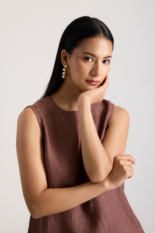 A woman with long black hair posing in a sleeveless, wearing a whiter pearl earring dusty rose-brown linen top against a light gray background.