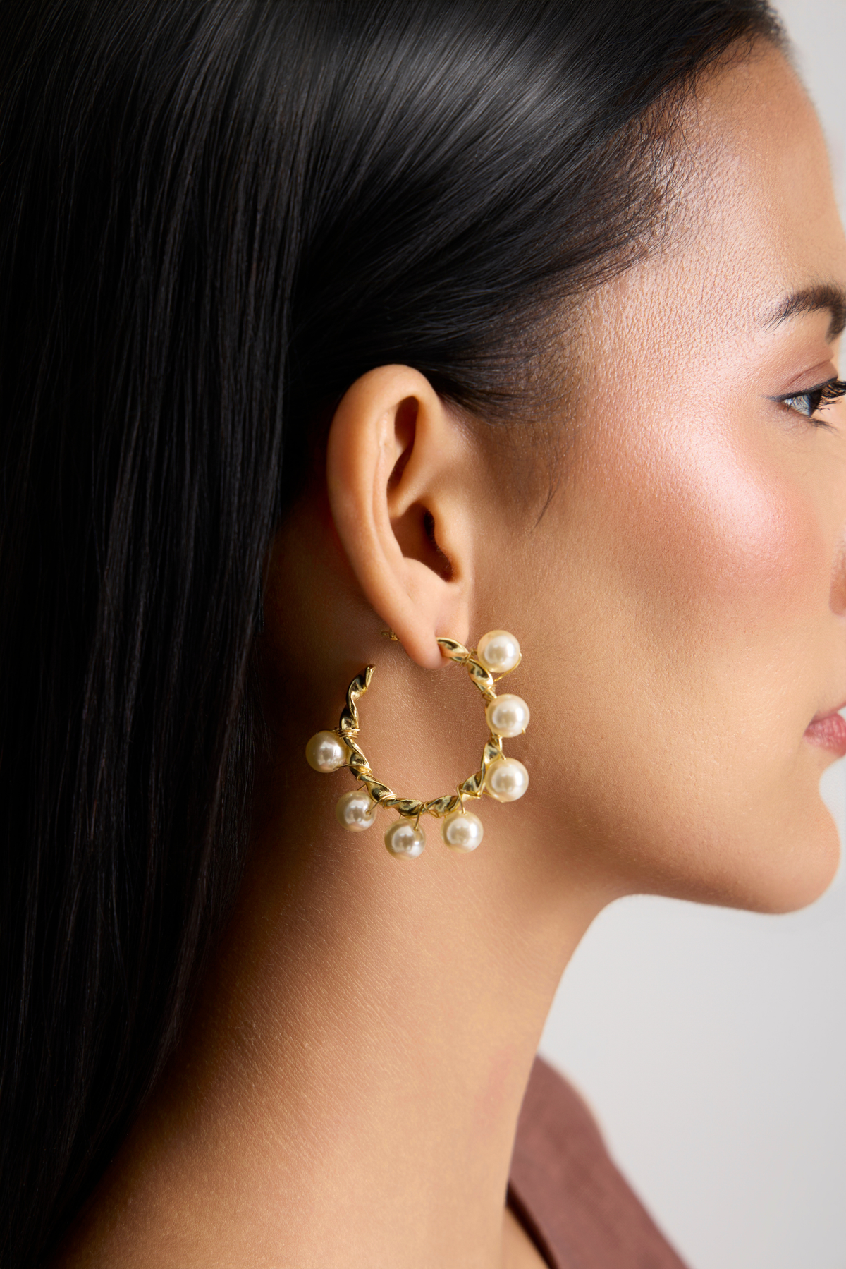 A close-up side profile of a woman wearing a gold hoop earring adorned with small white pearls, set against a clean light gray background.