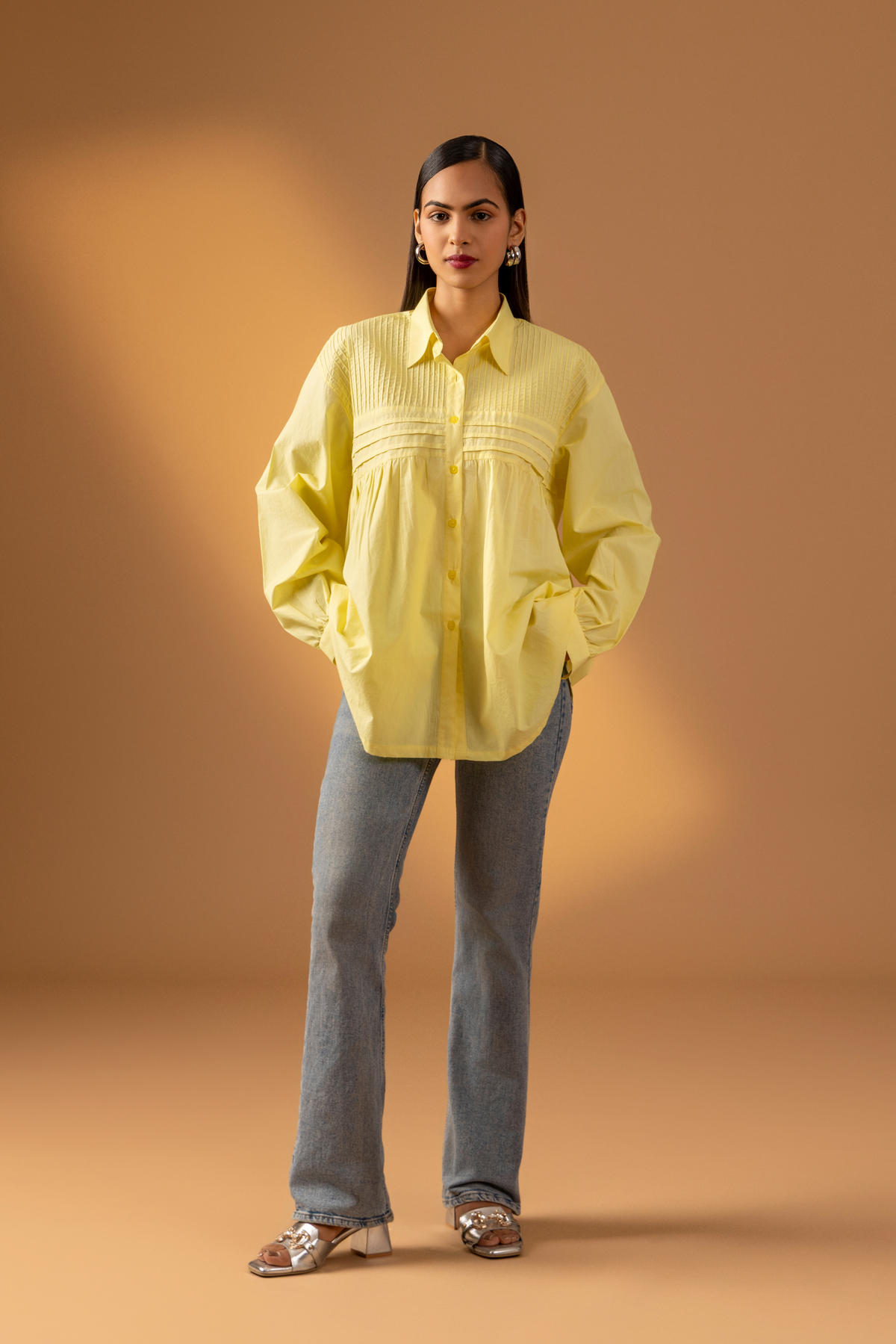 A woman wearing a stylish, oversized yellow shirt with delicate pleating, paired with light denim jeans, set against a warm backdrop.