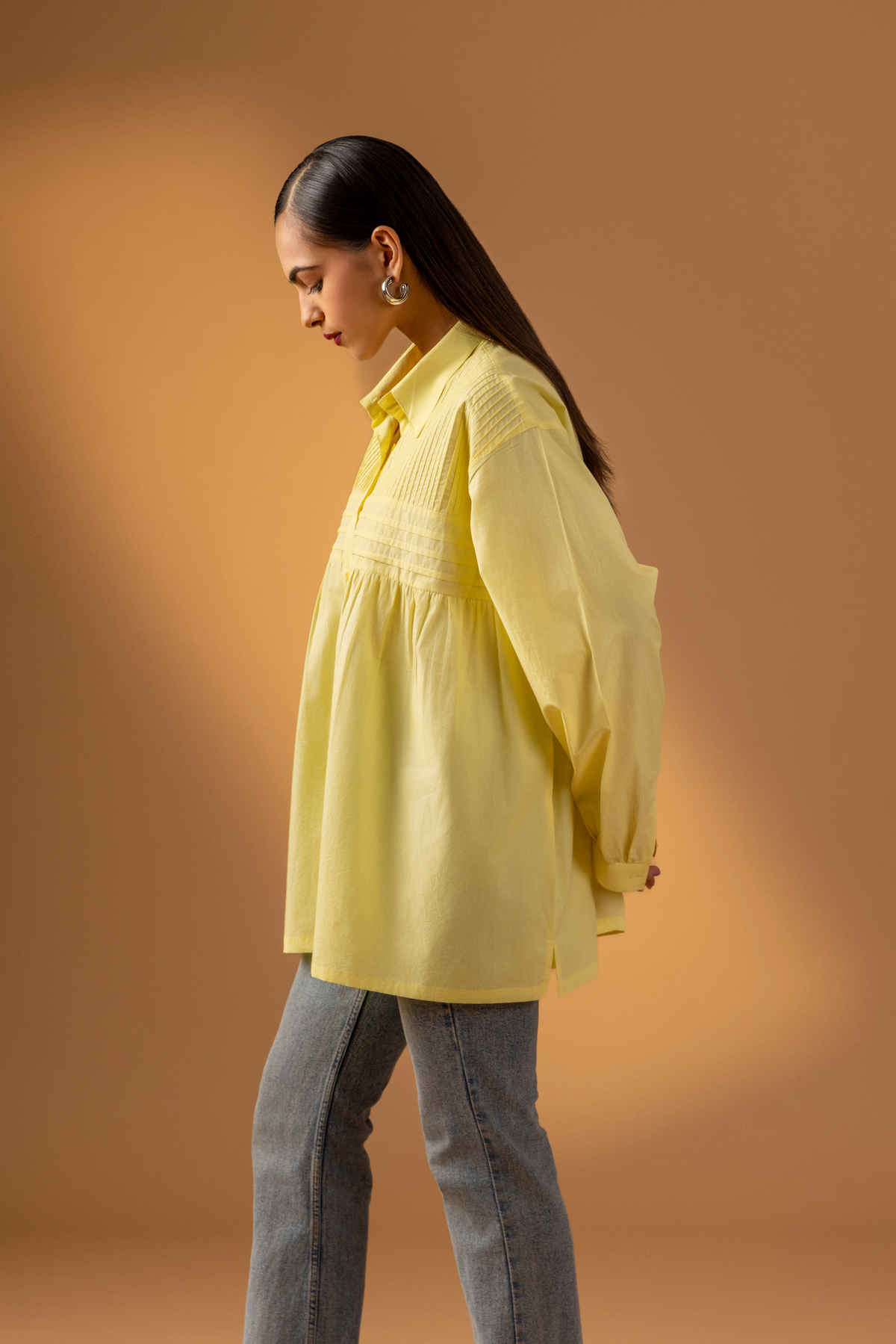 Side view of a woman wearing a stylish, oversized yellow shirt with delicate pleating, paired with light denim jeans, set against a warm backdrop.