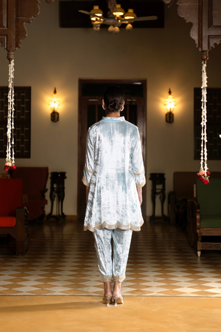 Back view of a woman wears a light blue ethnic velvet co-ord set outfit, standing gracefully in a spacious, elegantly decorated room with warm lighting and traditional furniture.