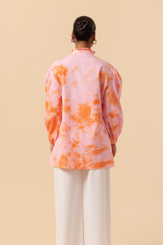 Back view of a model wears a vibrant light pink & orange tie-dye shirt dress with puffed sleeves, paired with white wide-leg trousers and stylish sandals.
