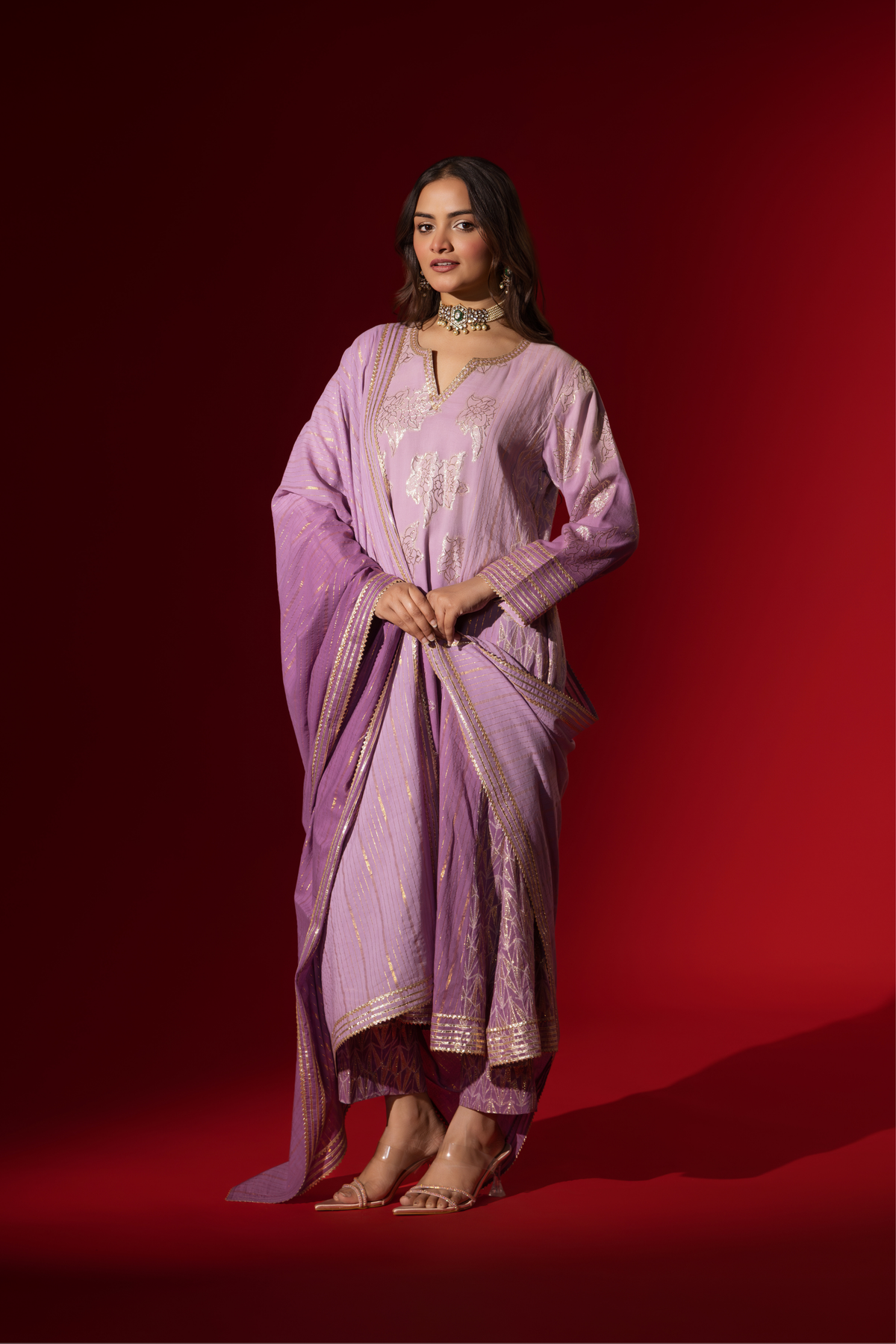 Side tilt view of a woman showcases an elegant lilac anarkali kurta set with intricate designs, contrasting beautifully against a deep red background.