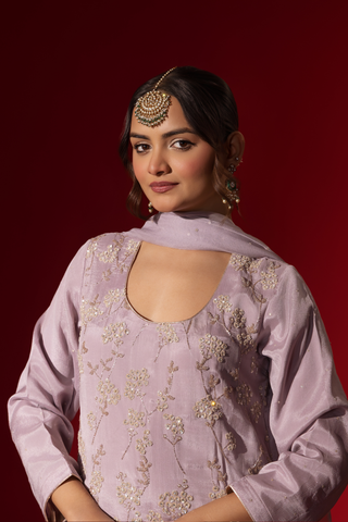 Closeup view of a woman dressed in a Lilac Radiance Embroidered Kurta Set stands confidently against a deep red background. Elegant traditional attire.