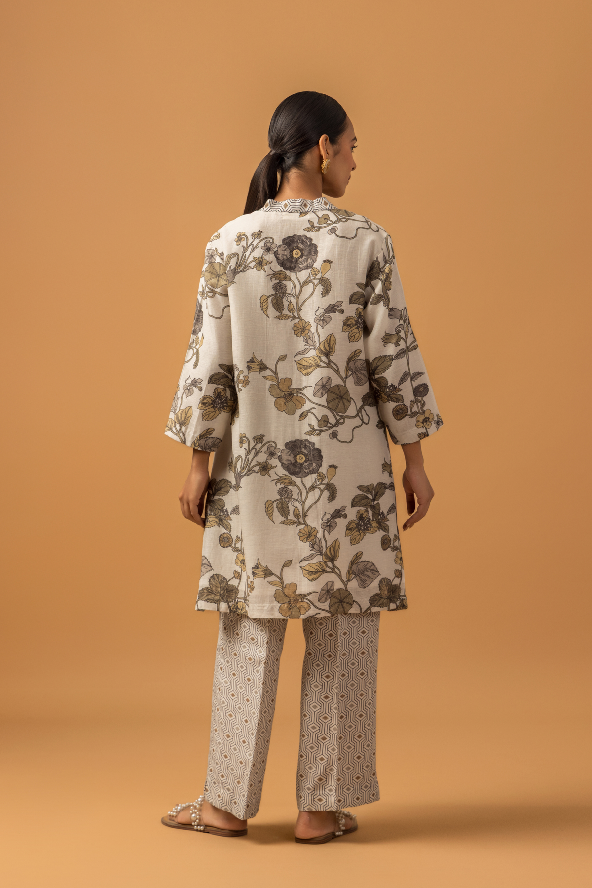 Back view of a woman stands against a warm tan background wearing a loose, floral-patterned co-ord set over patterned pants. The tone is calm and minimalist, with neutral colors 