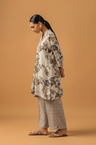 Side view of a woman stands against a warm tan background wearing a loose, floral-patterned co-ord set over patterned pants. The tone is calm and minimalist, with neutral colors 