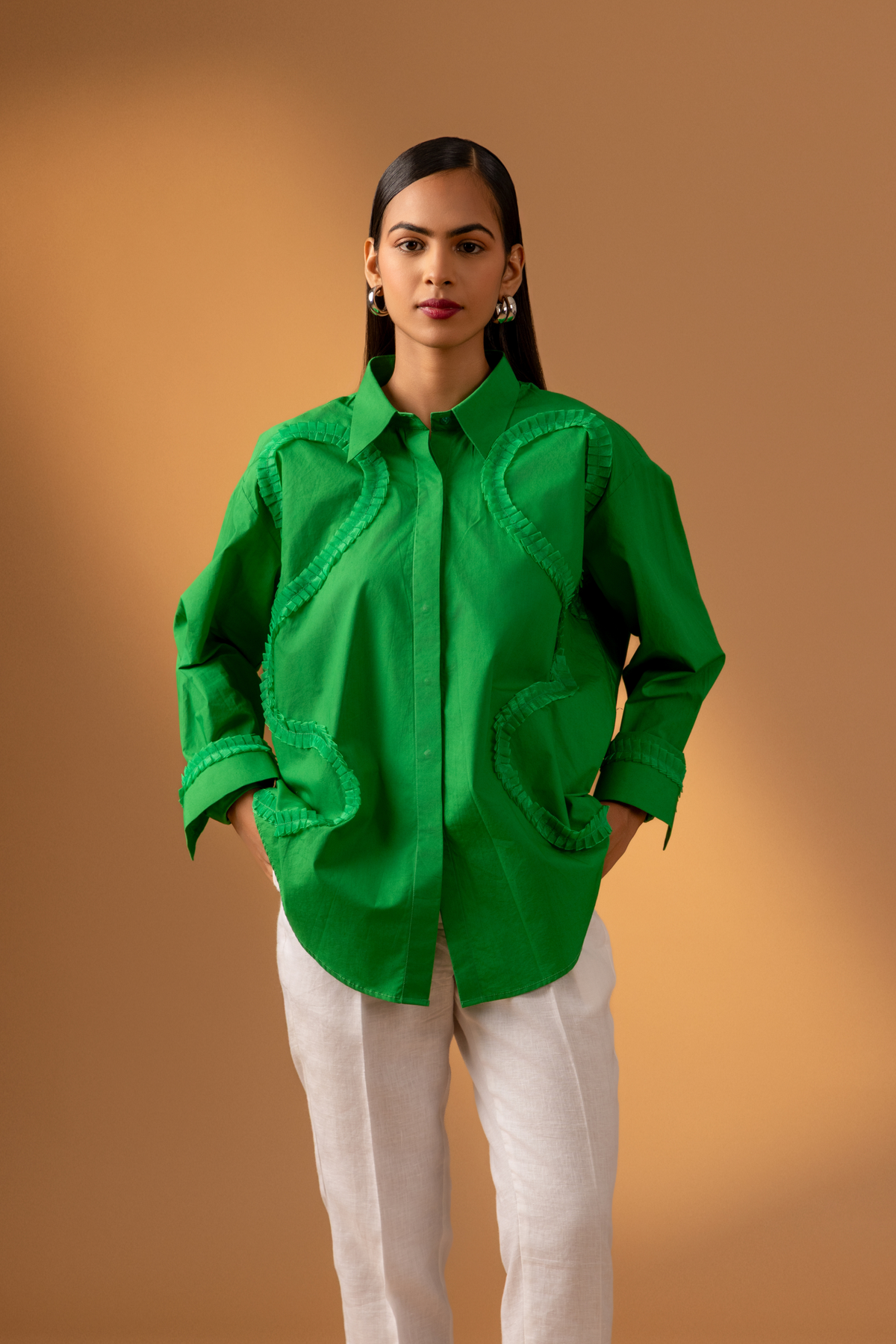 A model wears a vibrant green shirt with unique detailing, paired with white pants & hands in pocket, against a soft brown background.