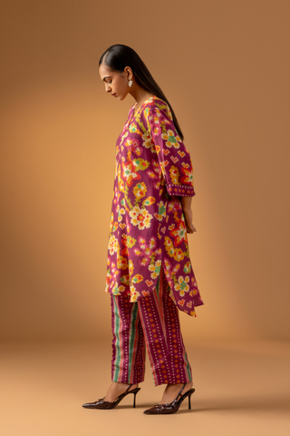 Side view of a model showcases a magenta floral-patterned co-ord set paired with colorful striped pants against a warm, neutral background.