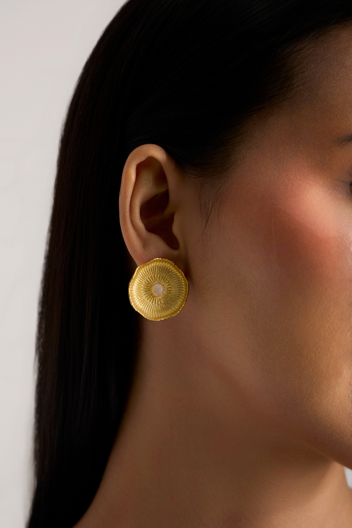A close-up side profile of a woman wearing a textured gold circular earring with a small white center, set against a plain light background.