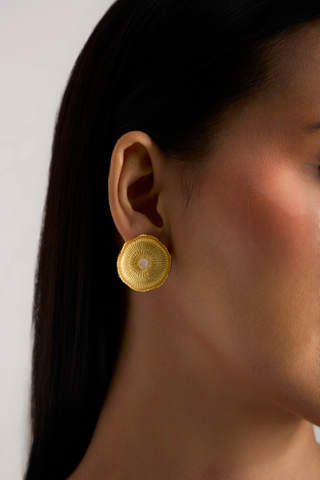 A close-up side profile of a woman wearing a textured gold circular earring with a small white center, set against a plain light background.
