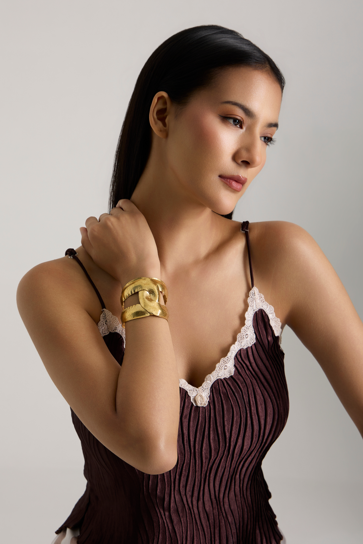 A woman wearing a chunky gold cuff bracelet with a central round charm, styled with a dark pleated camisole top, against a neutral gray studio background.