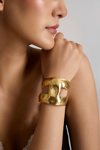 A close-up lifestyle shot of a model wearing a chunky, textured gold cuff bracelet on her wrist, set against a soft neutral gray background.