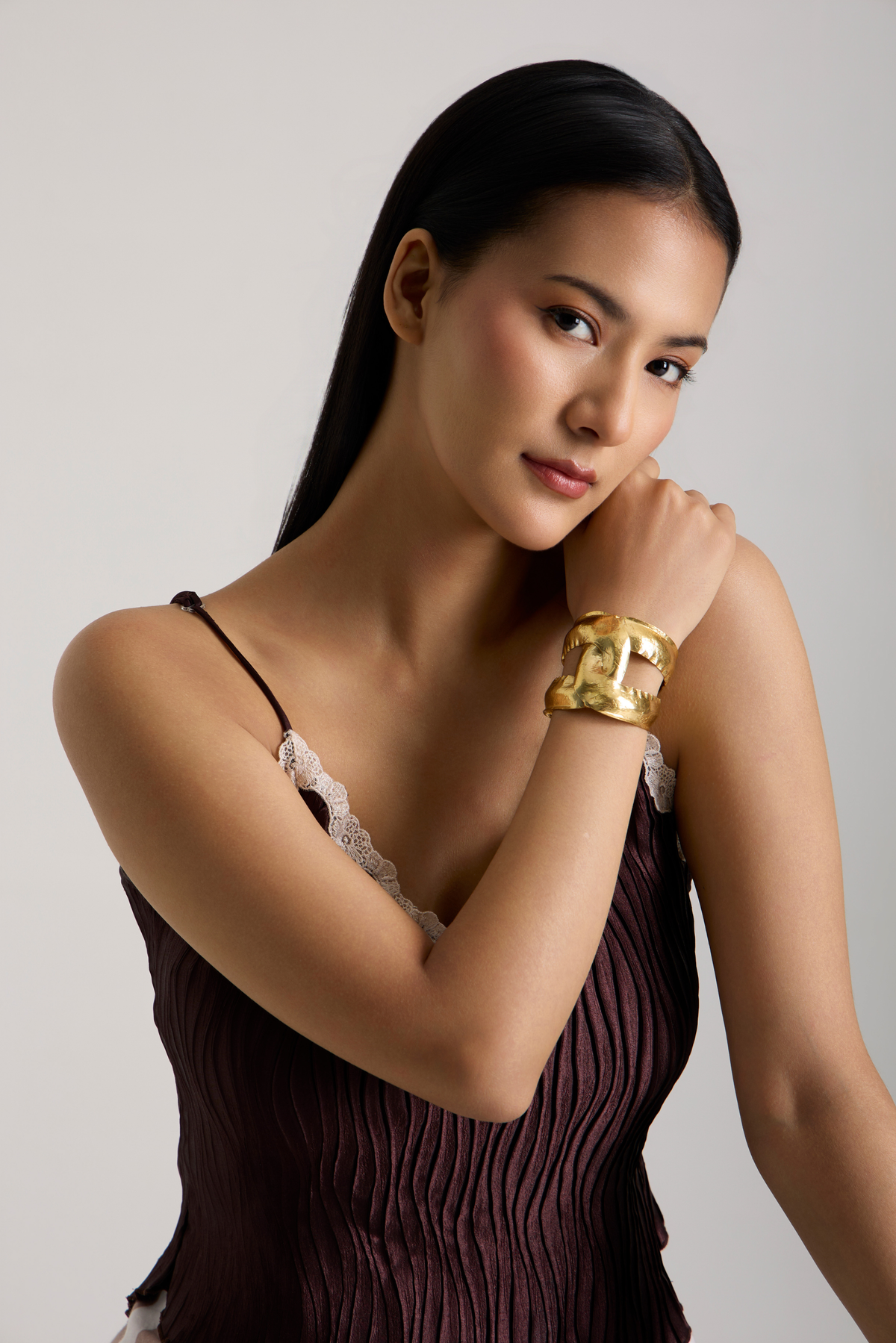 A woman wearing a chunky gold cuff bracelet, posing in a dark pleated camisole dress against a light gray background.