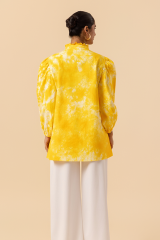 Back view of a woman wears a vibrant yellow tie-dye shirt dress with puffed sleeves, paired with white wide-leg trousers and stylish sandals.