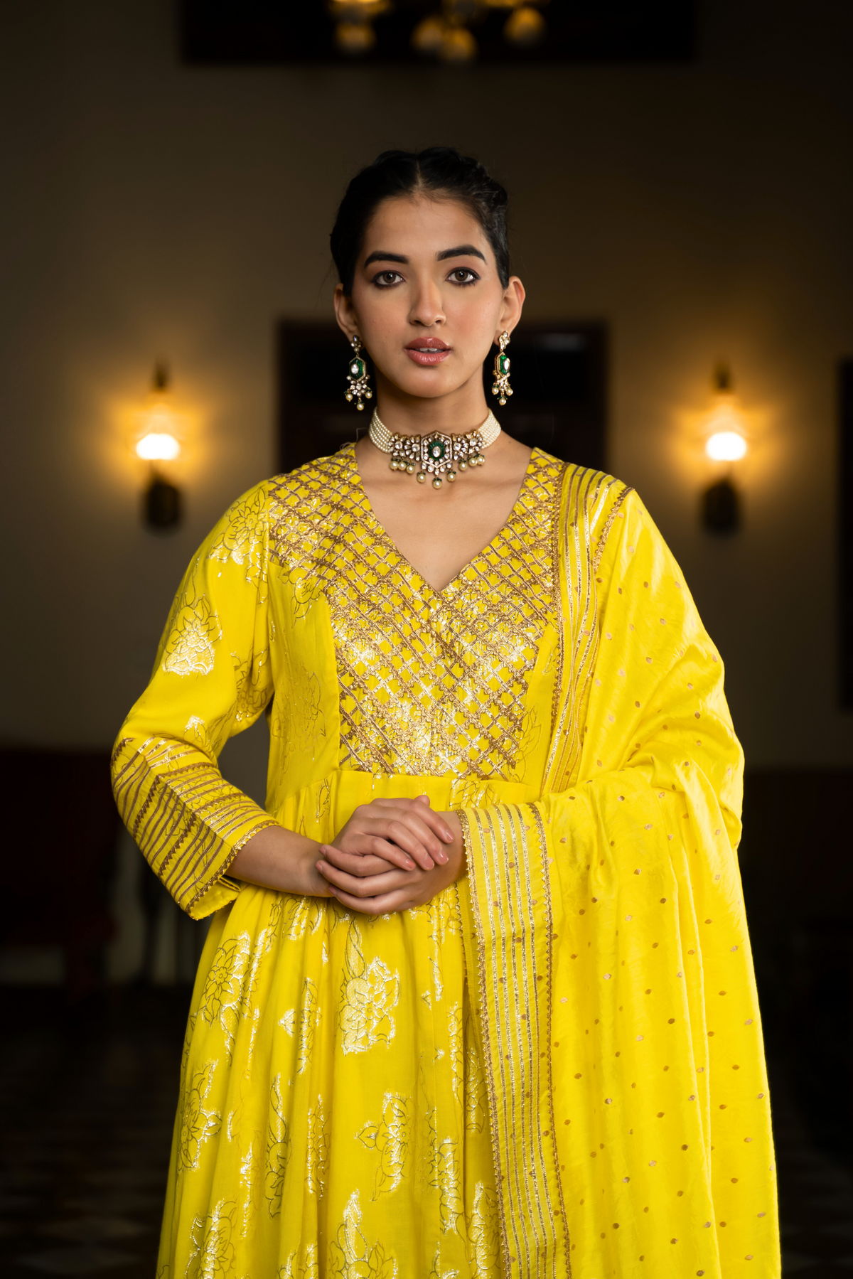 Closeup view of a  woman in a yellow anarkali kurta with intricate gold patterns with matching pants and dupattaa, standing in a beautifully decorated interior.