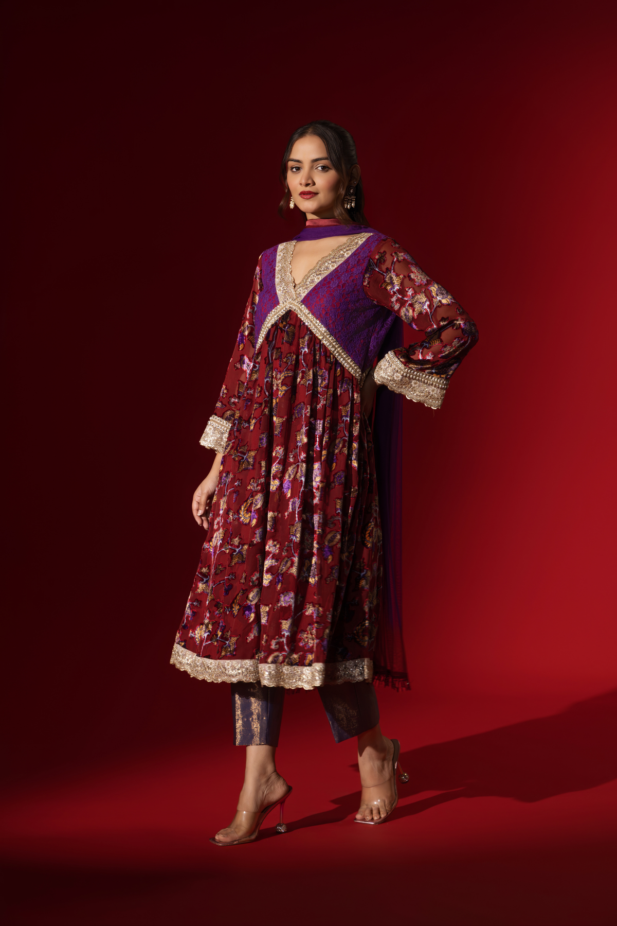 Side tilt view of a model poses in a vibrant maroon floral velvet anarkali kurta set with purple accents and a golden trim, set against a rich red background.
