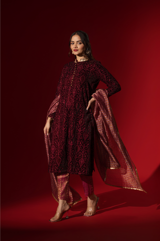 Side view of a person stands confidently in a rich, red traditional outfit adorned with intricate patterns and a shimmering gold-accented shawl against a red backdrop.
