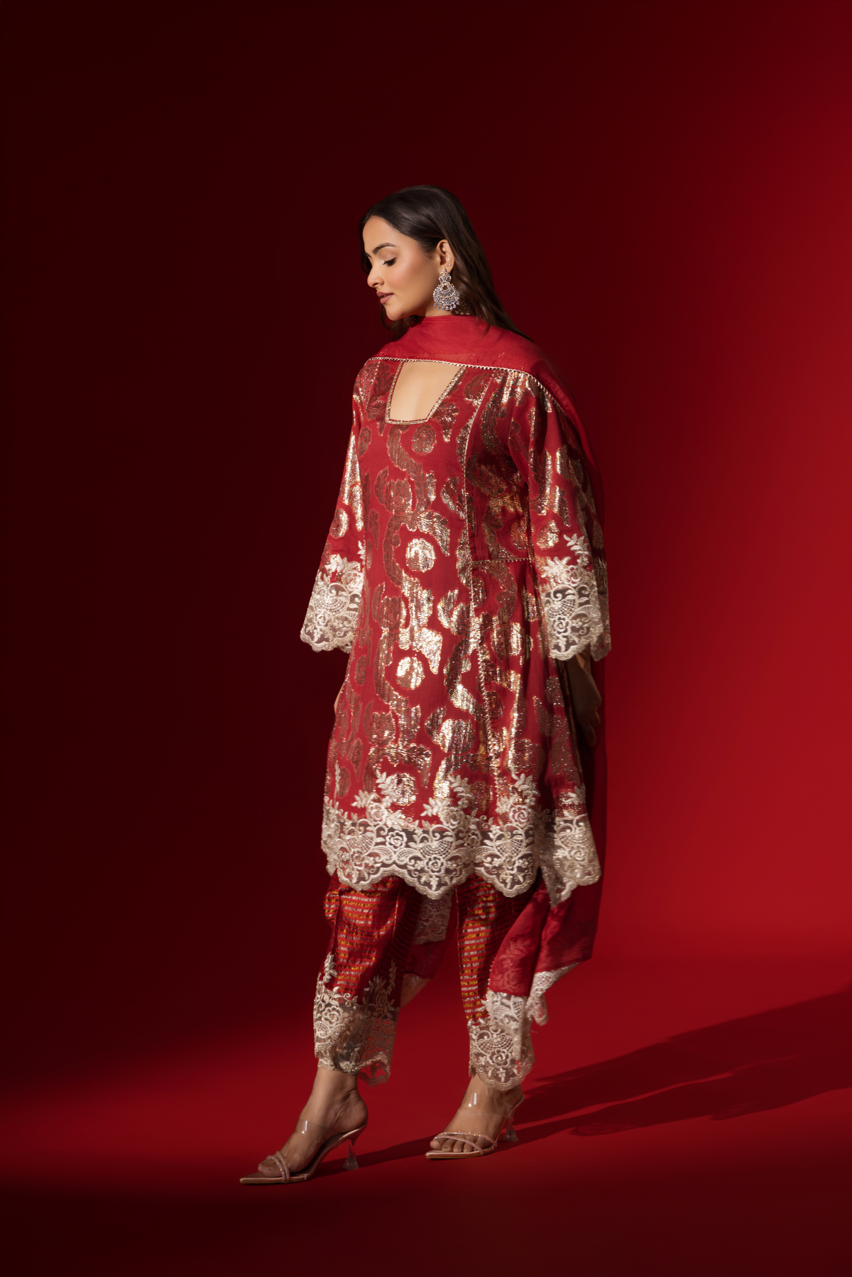 Side view of a woman poses in a maroon ethnic  shimmer kurta set outfit with intricate white embroidery against a vibrant red backdrop.