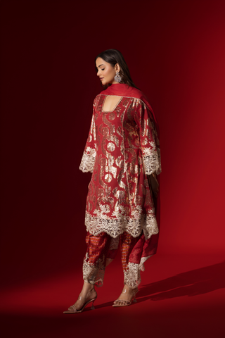 Side view of a woman poses in a maroon ethnic  shimmer kurta set outfit with intricate white embroidery against a vibrant red backdrop.
