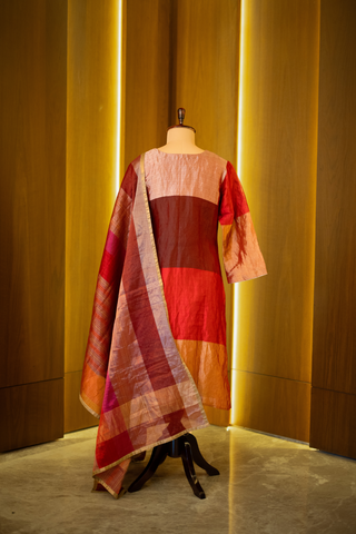 back view of multicolored patchwork kurta in shades of red, pink, and gold, paired with a coordinating striped dupatta, displayed on a mannequin against a warm wooden backdrop.