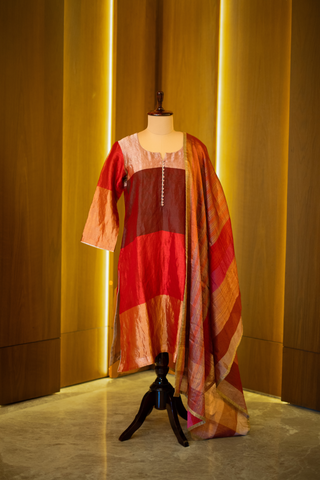 multicolored patchwork kurta in shades of red, pink, and gold, paired with a coordinating striped dupatta, displayed on a mannequin against a warm wooden backdrop.