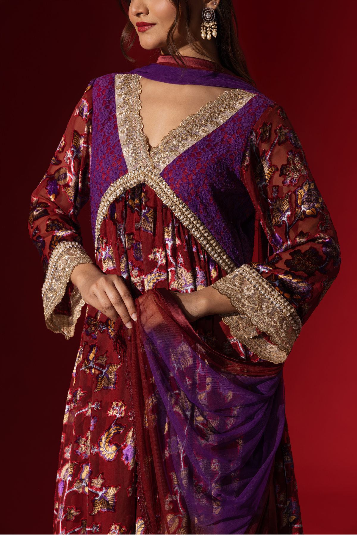 Closeup view of a model's hand in a vibrant maroon floral velvet anarkali kurta set with purple accents and a golden trim, set against a rich red background.