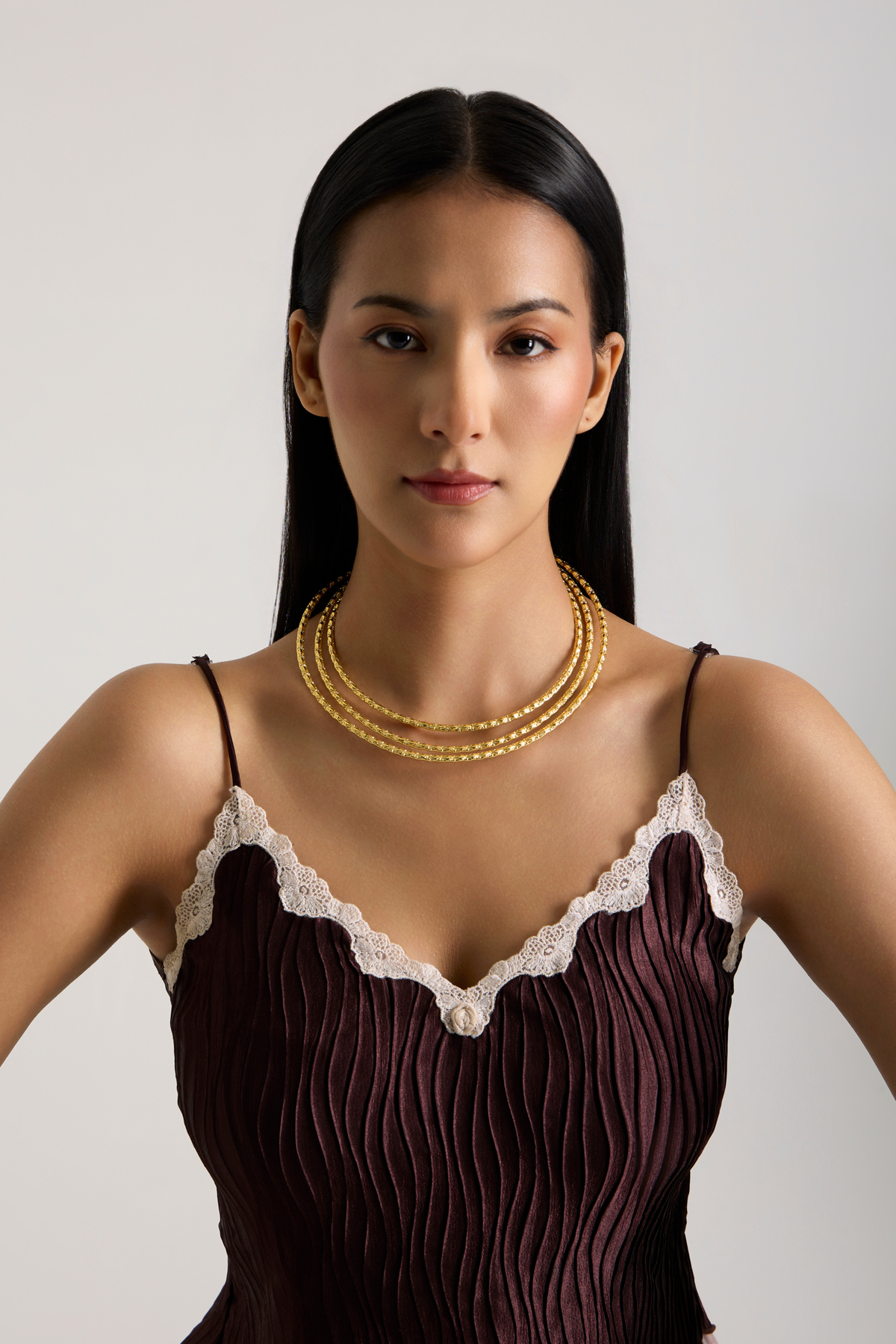 A woman with long black hair is shown from the chest up wearing a dark plum pleated camisole with white lace trim and a multi-strand gold necklace, against a light gray studio background.