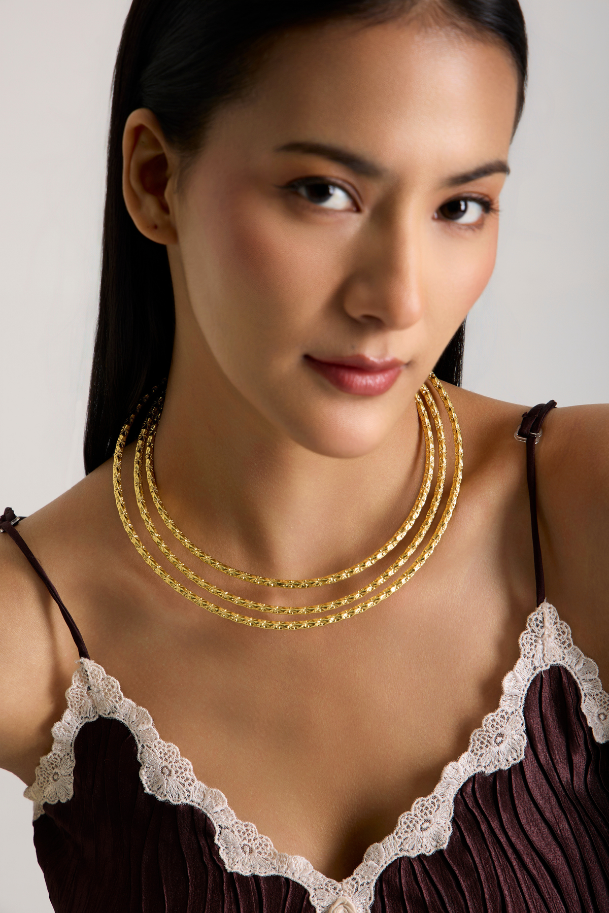 A close-up of a woman wearing a gold three-strand beaded necklace, styled with a dark brown lace-trimmed camisole against a light gray background.
