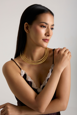 A woman with long dark hair models a gold multi-strand beaded necklace. She is wearing a black camisole with white lace trim, posed against a neutral light gray background.