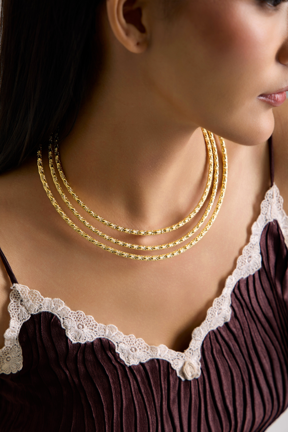 A close-up of a model wearing a multi-strand gold chain necklace with textured links, styled with a dark pleated top featuring white lace trim.