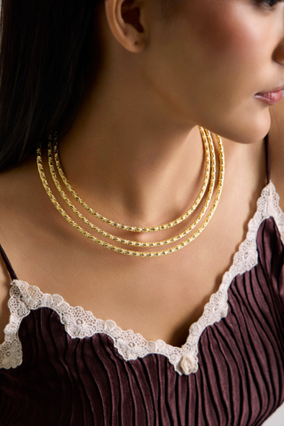 A close-up of a model wearing a multi-strand gold chain necklace with textured links, styled with a dark pleated top featuring white lace trim.