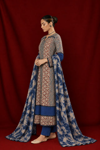 A woman modeling a traditional ajrakh anarkali kurta set  with a long dupatta, featuring a beige kurta with red and blue geometric patterns, solid blue pants, and a matching blue and beige patterned dupatta, set against a dark red draped backdrop.