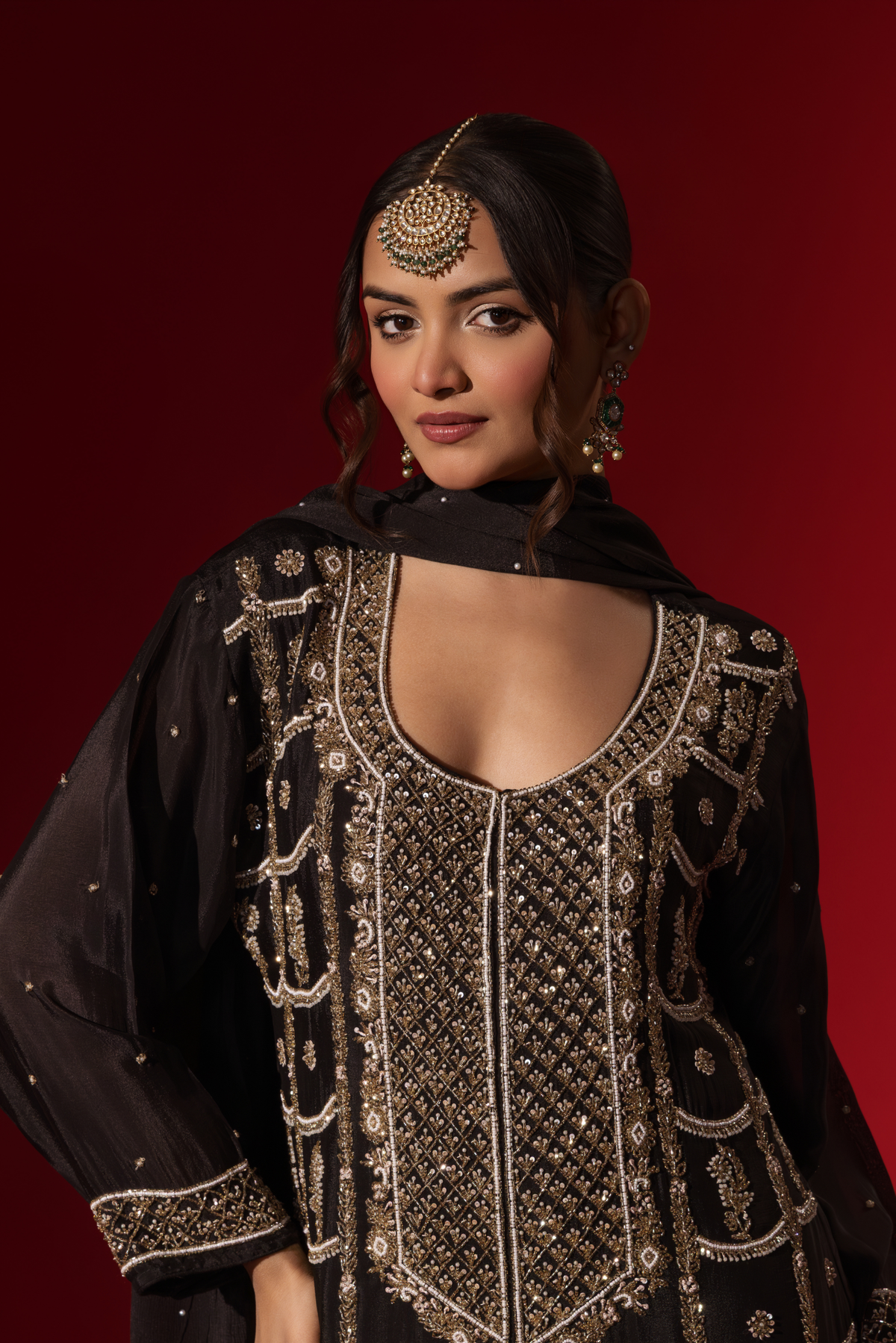 Closeup view of a woman showcases an elegant black embroidered kurta set with intricate gold embroidery against a rich red backdrop.