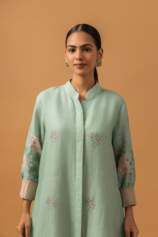 Closeup view of a woman in a pastel green, floral co-ord set with delicate embroidery stands against a beige background. She is wearing beige pants, sandals, and hoop earrings, conveying a serene and elegant tone.