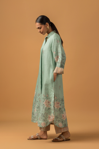 Side view of a woman in a pastel green, floral co-ord set with delicate embroidery stands against a beige background. She is wearing beige pants, sandals, and hoop earrings, conveying a serene and elegant tone.