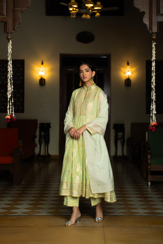 Side view of a woman in a mint green anarkali kurta set outfit stands elegantly in a beautifully decorated indoor space with warm lighting and classic furnishings.