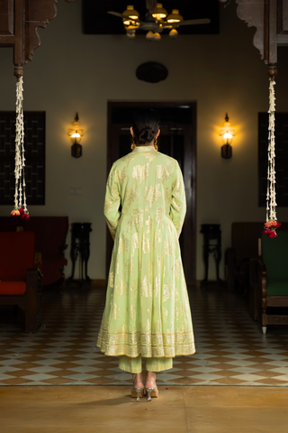 Back view of a woman in a mint green anarkali kurta set outfit stands elegantly in a beautifully decorated indoor space with warm lighting and classic furnishings.