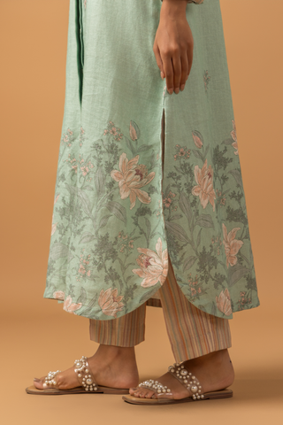 Closeup view of a woman in a pastel green, floral co-ord set with delicate embroidery stands against a beige background. She is wearing beige pants, sandals, and hoop earrings, conveying a serene and elegant tone.