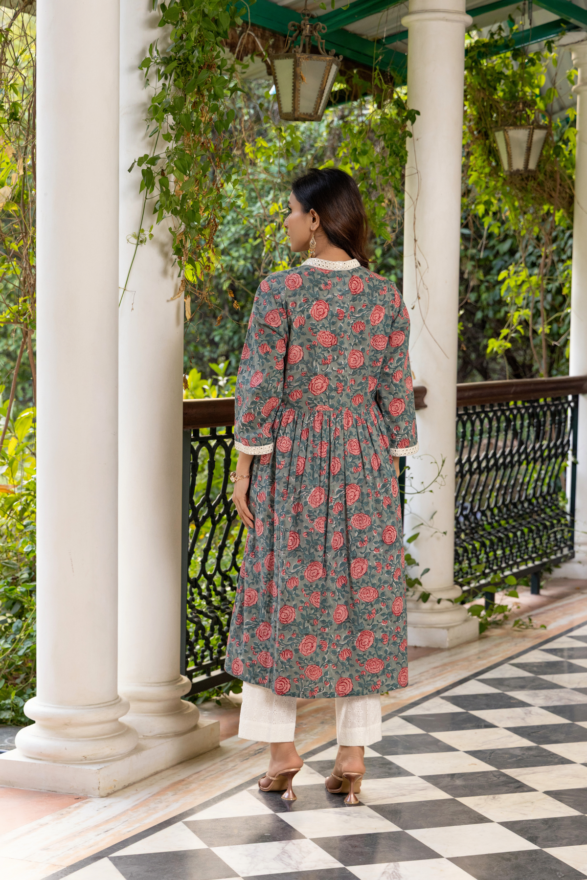 Back view of a woman stands elegantly on a black-and-white checkered floor, wearing a floral handblock printed cotton kurta set and white pants, surrounded by greenery. 